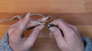 Hole den Faden über die Nadel mit der Häkelnadel und ziehe ihn durch die Schlaufe auf der Häkelnadel. Der Arbeitsfaden befindet sich jetzt vor der Stricknadel. maschen anschlagen