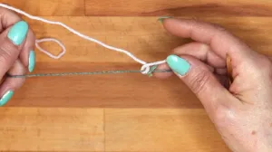 Lege den Arbeitsfaden um den Hilfsfaden und halte das lose Ende fest.. Maschen Anschlagen