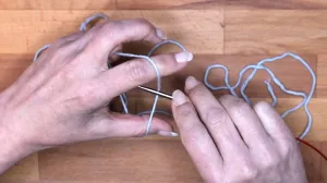 Lege den Faden vom Knäuel kommend über Zeigefinger und Daumen der linken Hand und führe die Nadel in die Lücke zwischen Faden und Hand.. Maschen Anschlagen